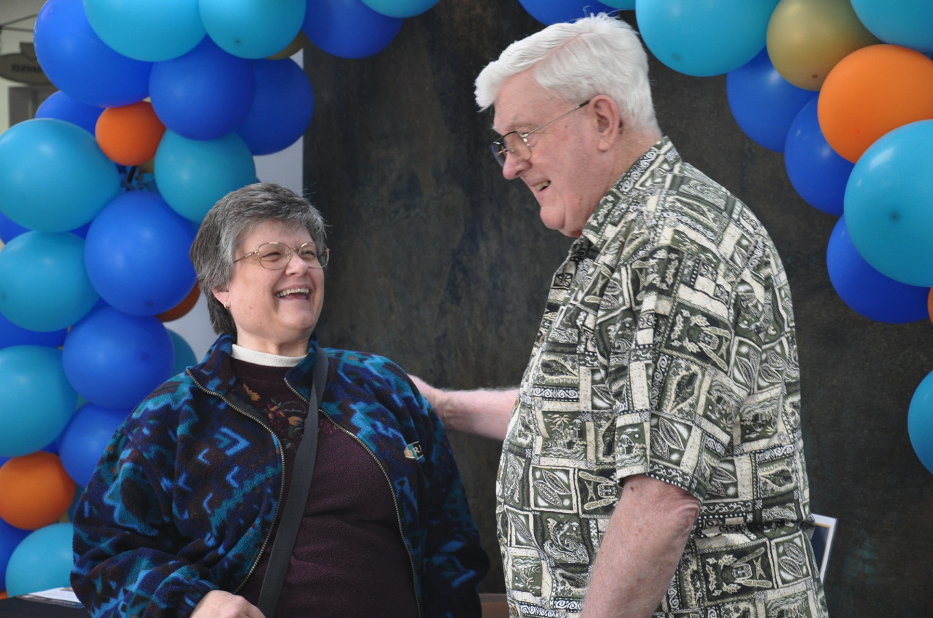 Two seniors, a man and a woman, laugh together under a balloon arch